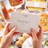 Hands holding a card with 'angel' and 'thinking of you' text over a table of assorted pastries and treats.